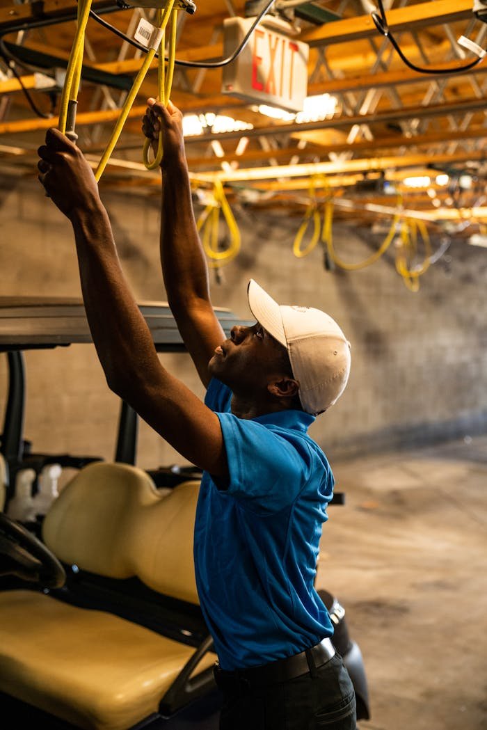 A man wearing a cap works on wiring above a golf cart in a garage.