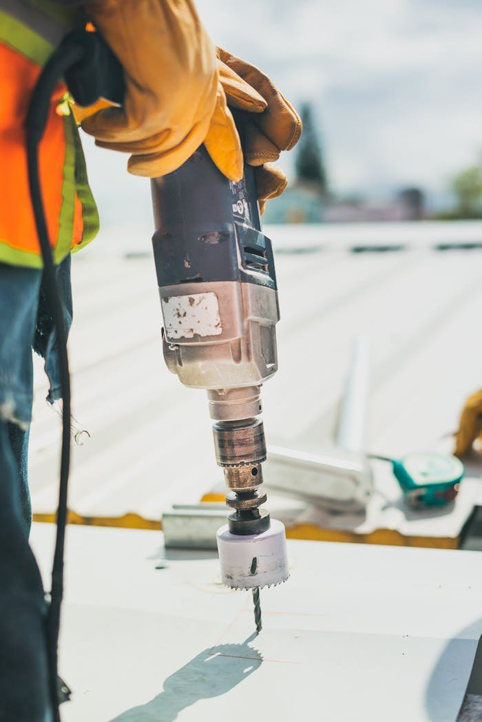 Worker drilling metal outdoors with a power drill for installation purposes. Safety gear evident.