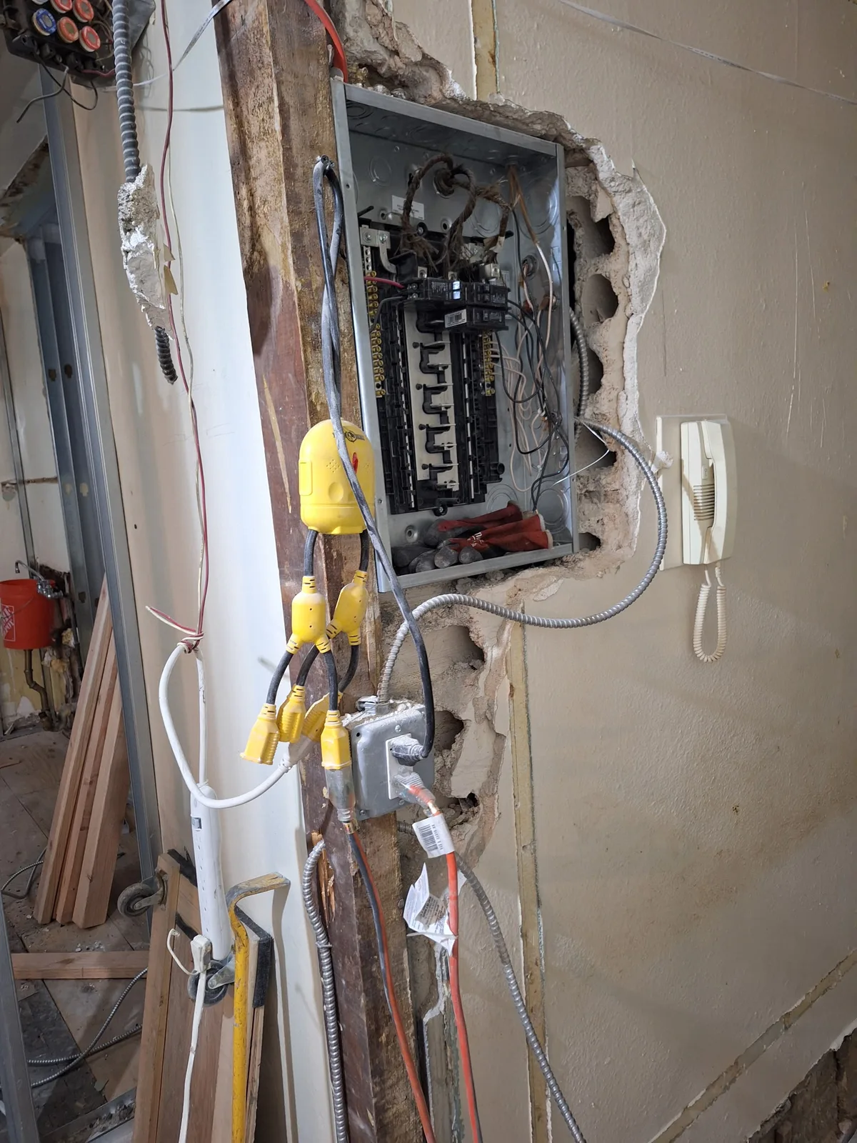 Open electrical panel in a Brooklyn house with the wall cut back, old cloth-covered conductors entering the top and temporary cord splitters powering the job